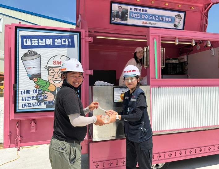 SM스틸 건설부문의 경기 용인시 양지지구 공동주택 신축공사 현장 근로자들이 지난달 25일 푸드트럭에서 팥빙수를 받으며 기념촬영을 하고 있다. 회사는 혹서기 근로자들의 건강 관리를 위해 온열질환 ‘제로(0)’ 캠페인을 운영하며 삼계탕, 팥빙수와 함께 보냉조끼, 넥쿨러 등 더위 대응 장비도 지원하고 있다. /사진제공=SM그룹